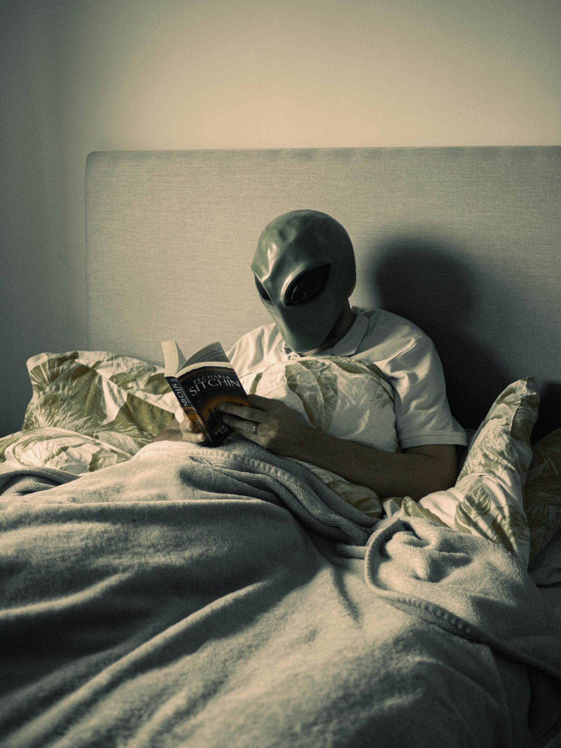 A person in an alien mask reading a book in bed.