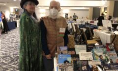 Shirley, wearing a cool hat and a peacock shawl, and Ira, wearing a mask and a snappy vest, in front of the vendors' table at Can*Con.