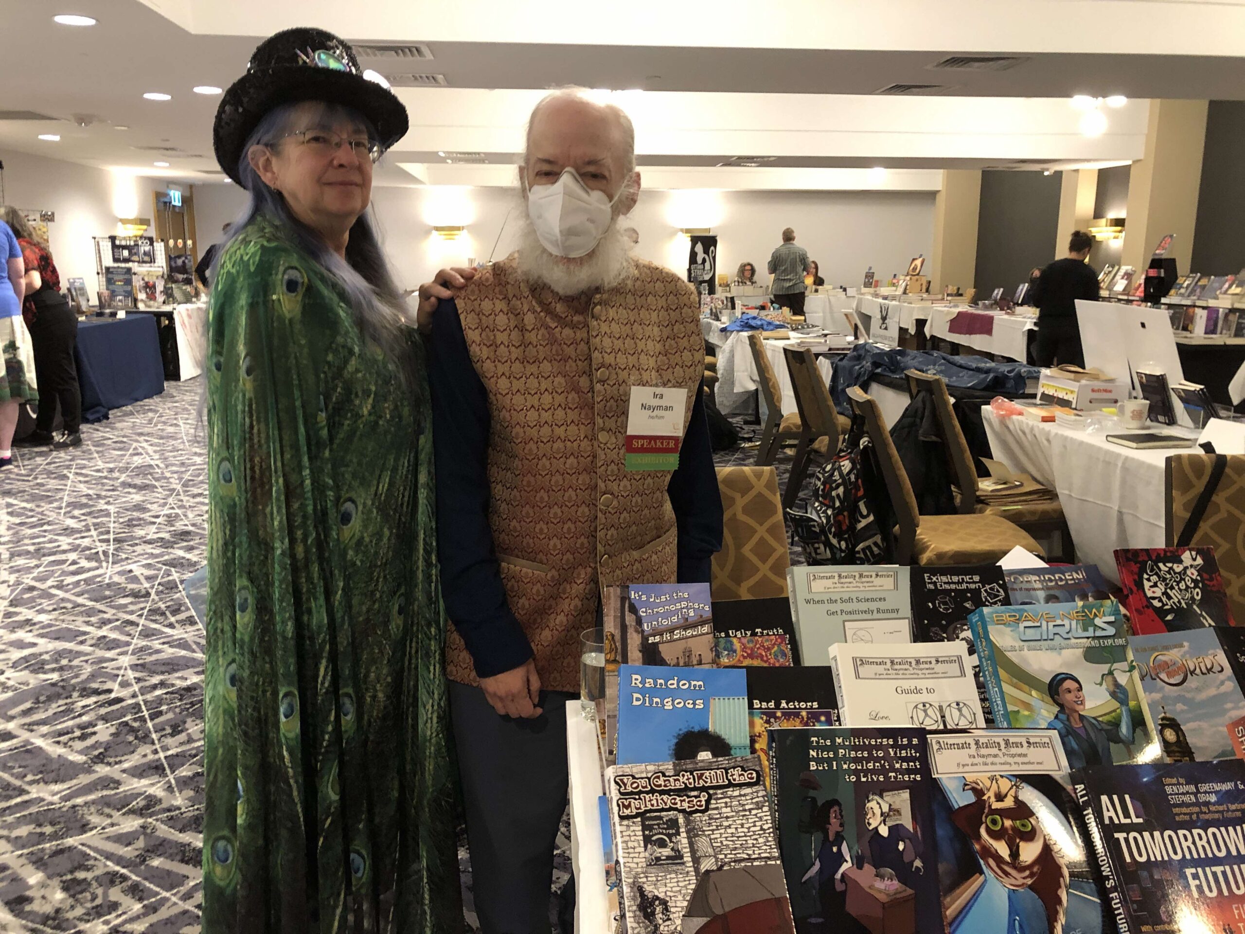 Shirley, wearing a cool hat and a peacock shawl, and Ira, wearing a mask and a snappy vest, in front of the vendors' table at Can*Con.